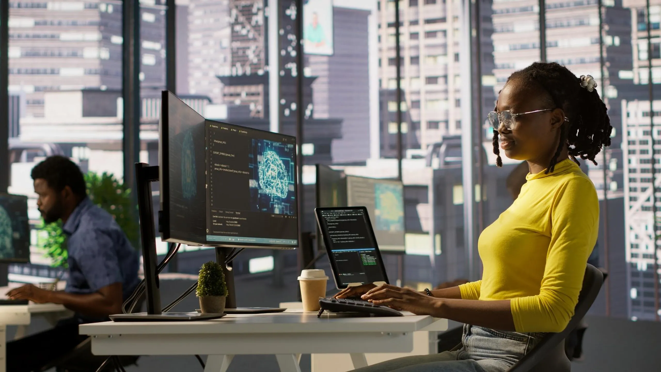 A woman in a yellow top and clear glasses types at a desk facing monitors that show code and a glowing brain graphic, while a colleague in a blue shirt works at the next desk, city towers visible through the windows behind them