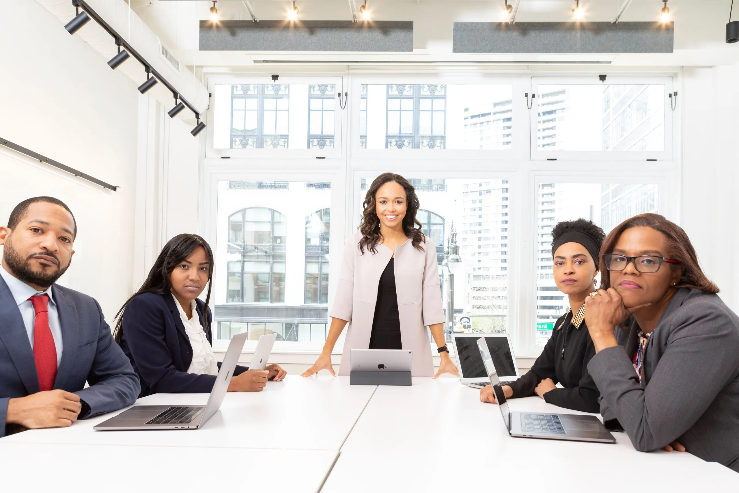 Five executives at a white boardroom table with laptops, a woman standing at its head and four seated colleagues looking on, tall windows onto city buildings behind them